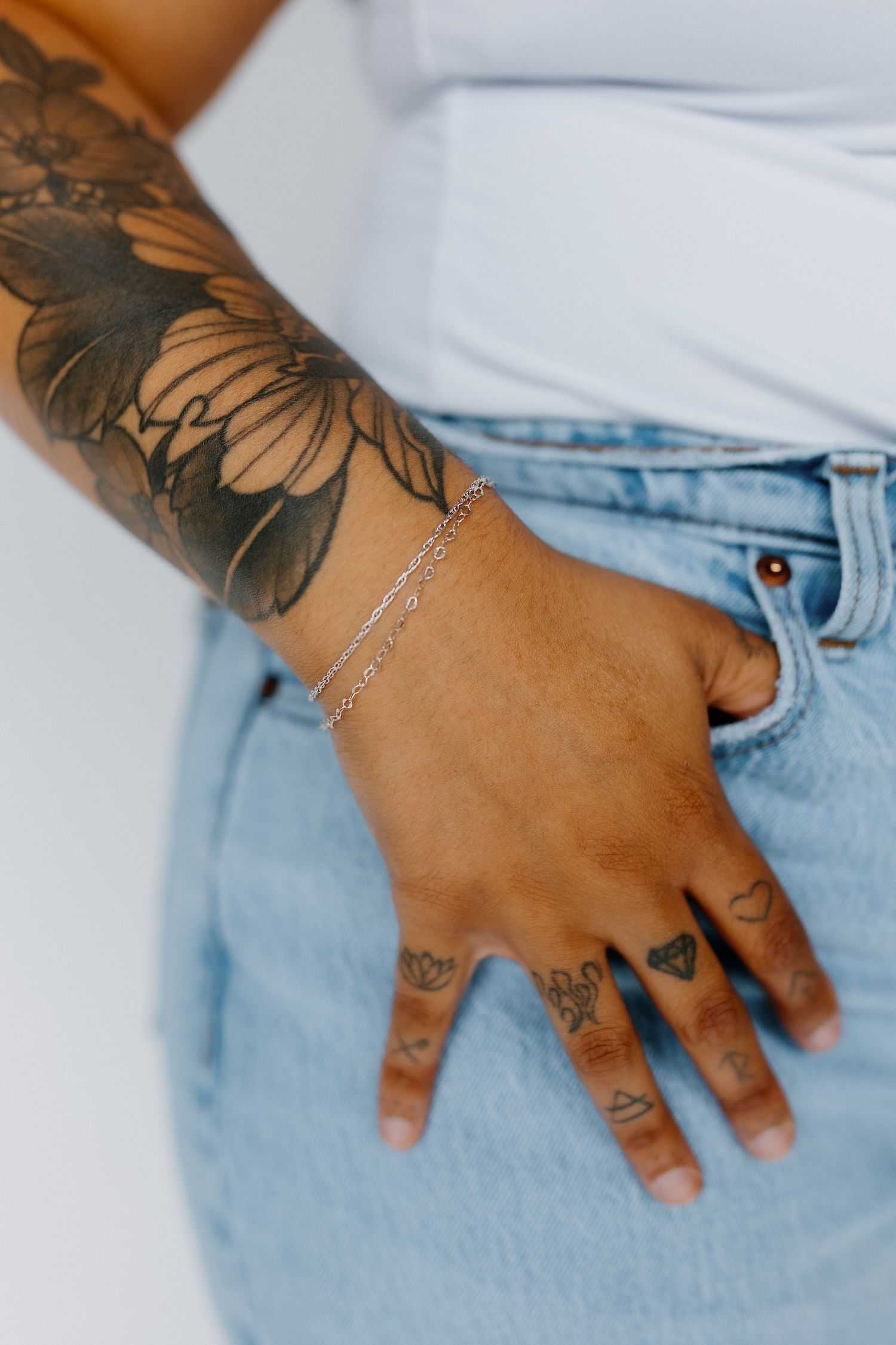 Person with floral tattoo on arm, wearing silver bracelet and jeans, hand in pocket.