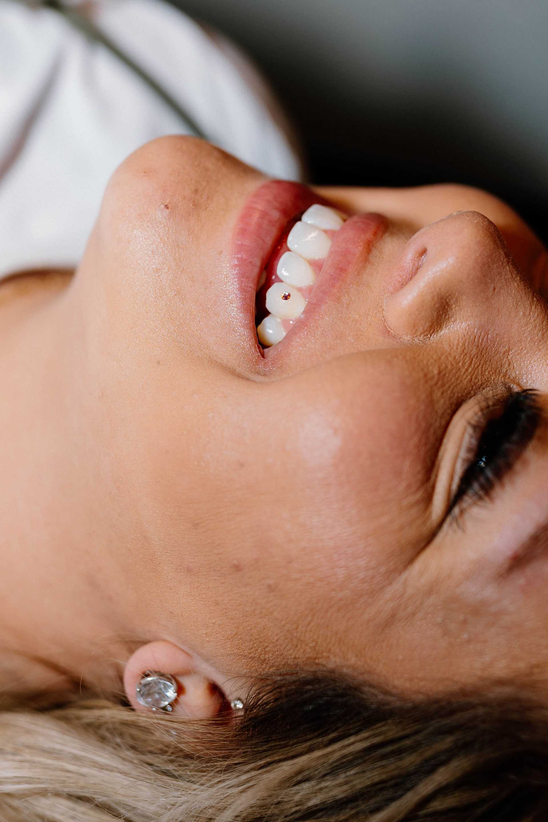 Close-up of a person smiling with a jewel in their visible ear and a beauty mark on their cheek.