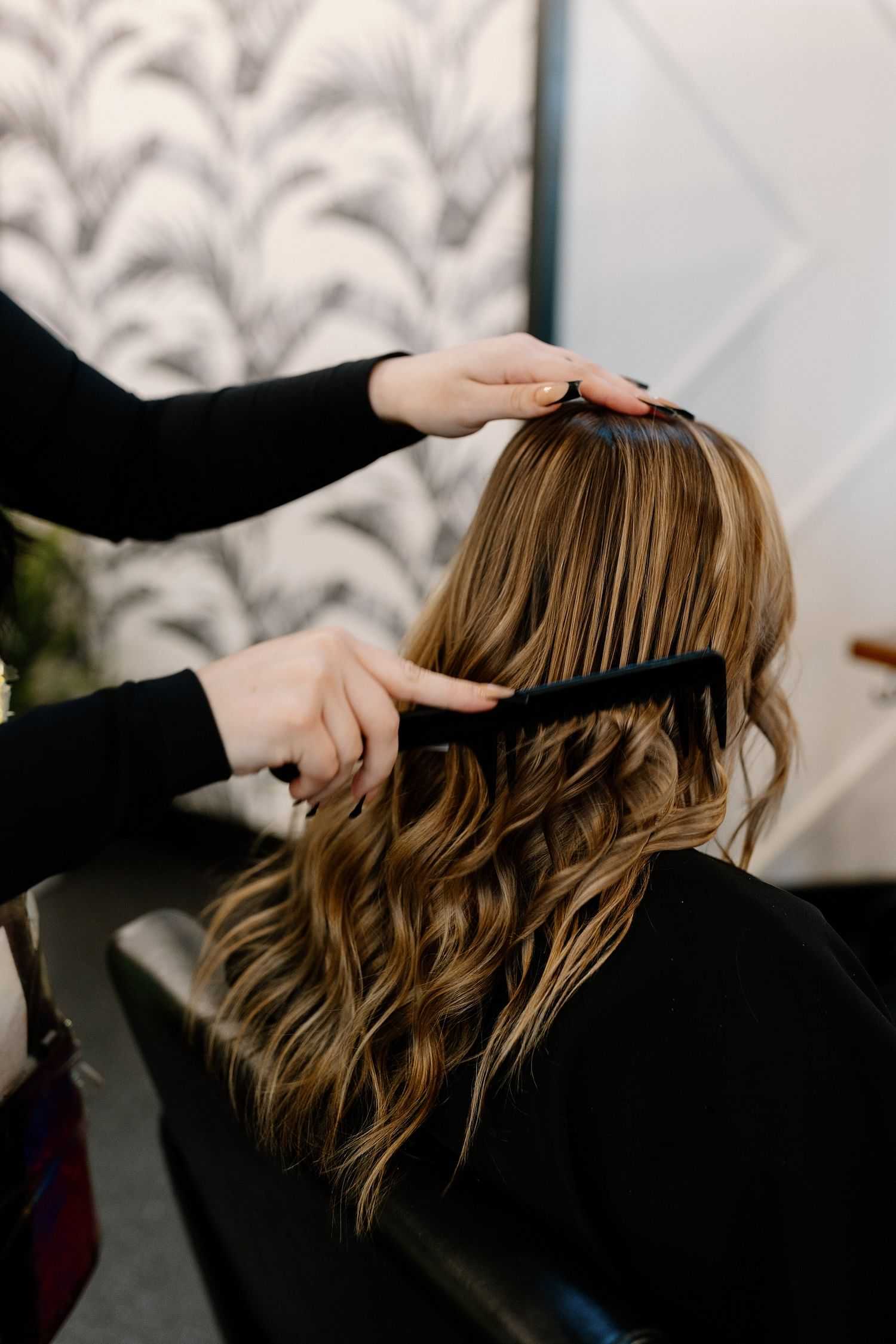 Stylist combing through wavy hair of a seated client in a salon.