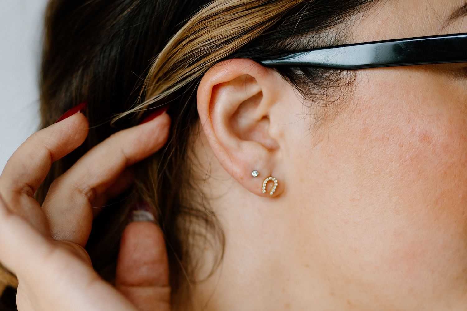 Woman wearing black glasses and a horseshoe-shaped earring touches the side of her head.